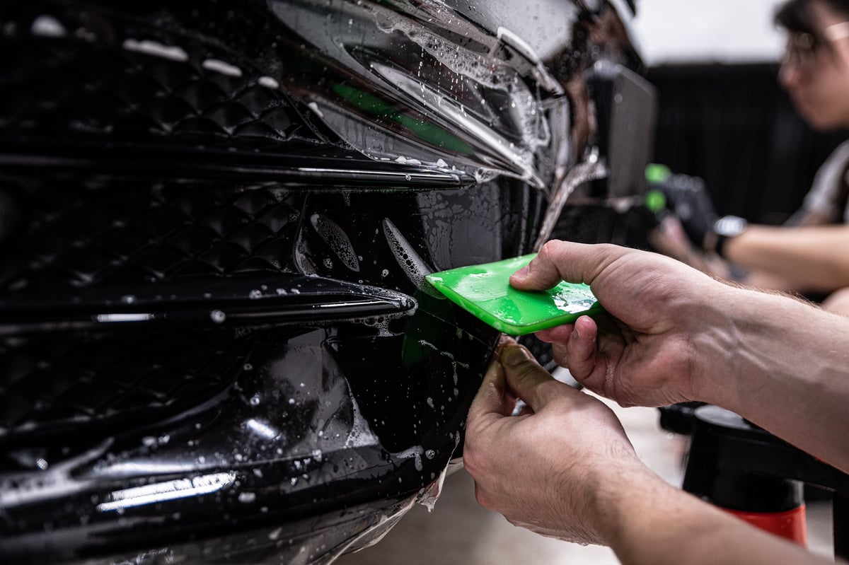 Technician applying a colourless protective PPF film to the bumper of a black car