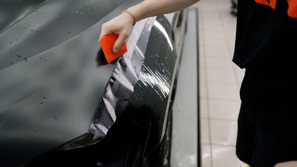 Automotive professional applying transparent protective vinyl film to a car hood with a squeegee